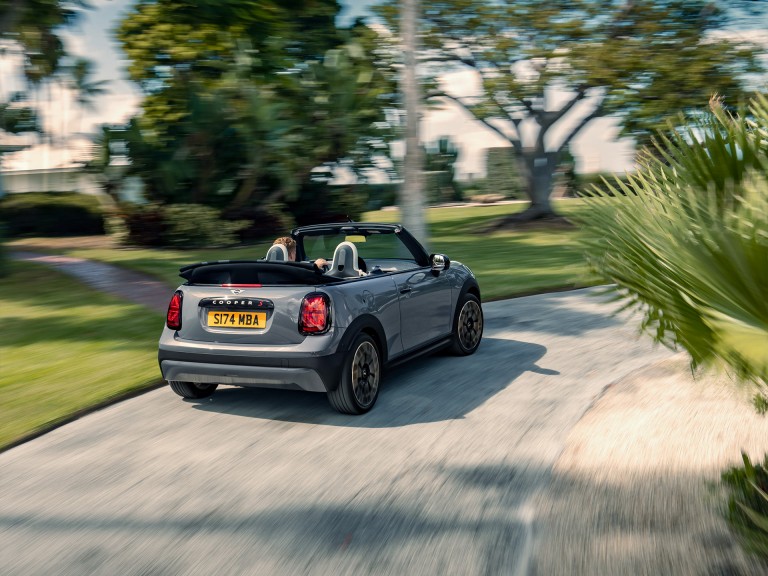 Angled back view of the new MINI Cooper Convertible in motion with the top down and a blurred, lush backdrop.
